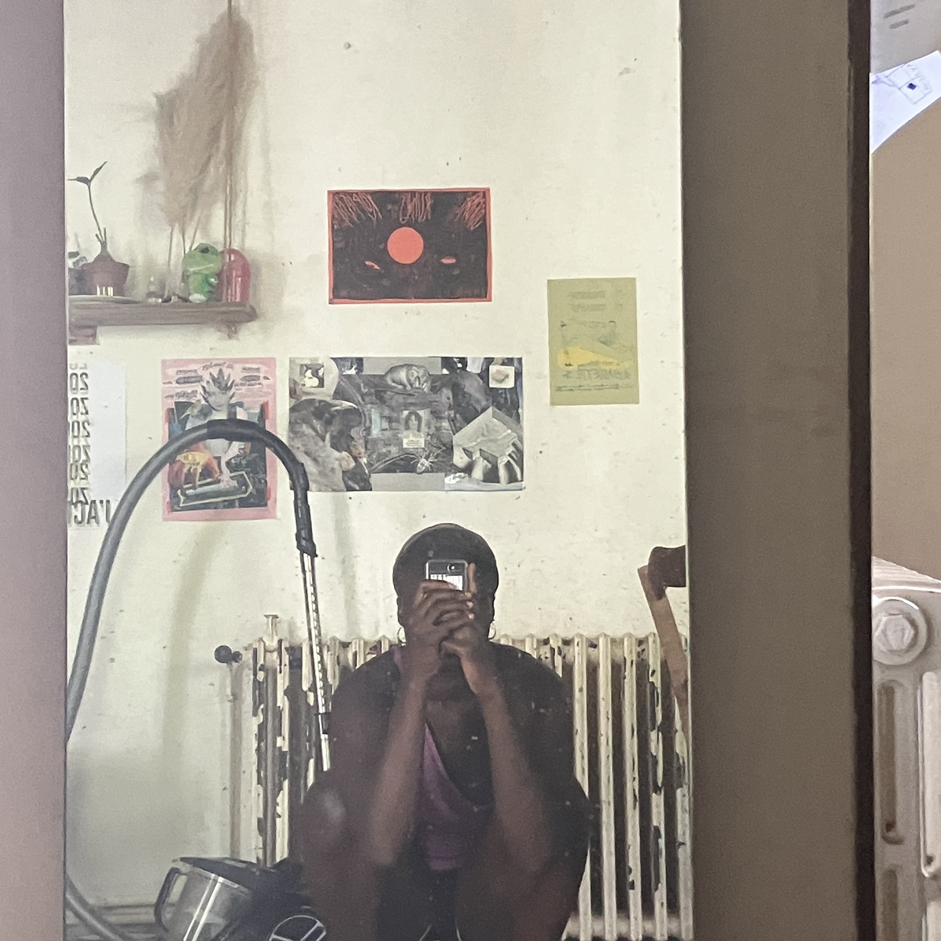 artist alero (a black non-binary person) takes a selfie in the mirror as they are squatting down on the floor with posters of various colours above their head